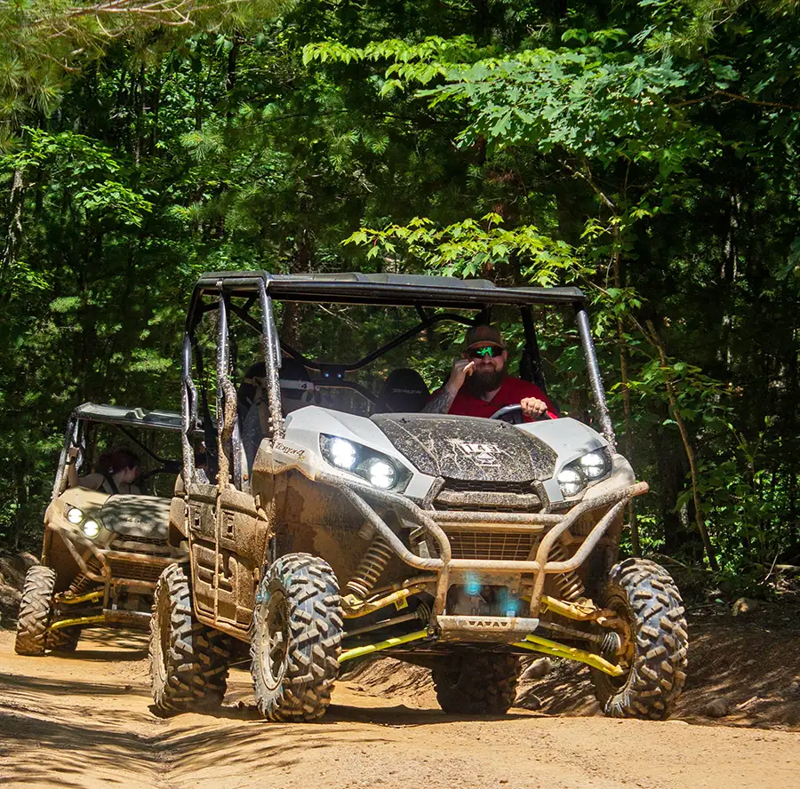 Two muddy UTVs driving through a forest on a guided off-road tour